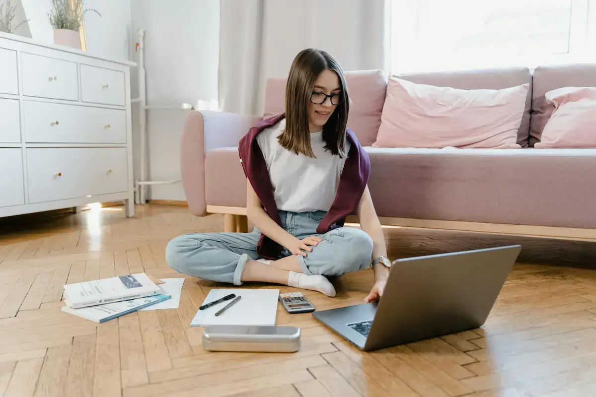 Young woman sitting cross-legged on wooden floor in a living room, using a laptop with study materials and pens around her.