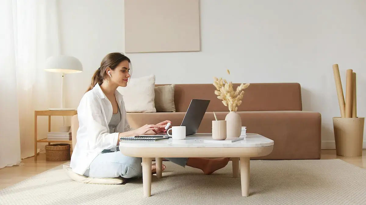 Woman sitting cross-legged on floor cushion working on a laptop at a low table in a cozy, neutral-toned living room.