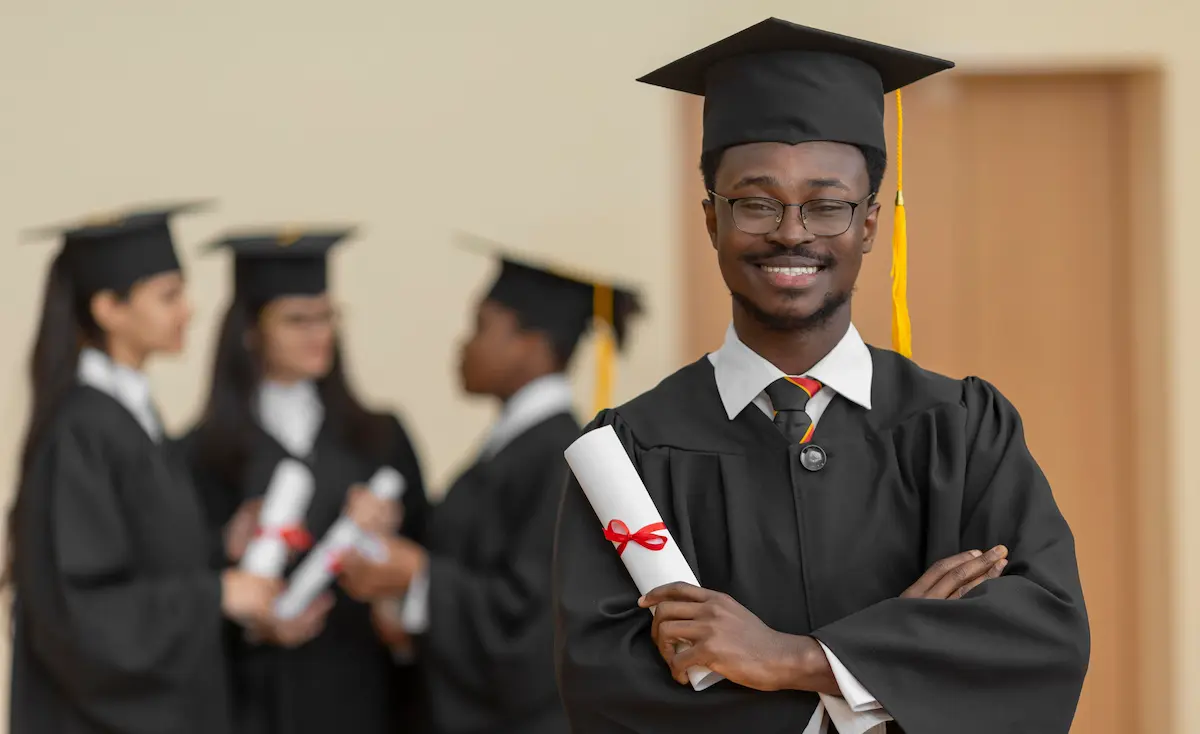 Smiling young graduate wearing glasses and holding a diploma with crossed arms, with three graduates in the background.