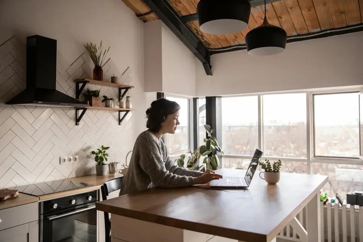Woman wearing headphones working on a laptop at a kitchen island with large windows and plants in the background.