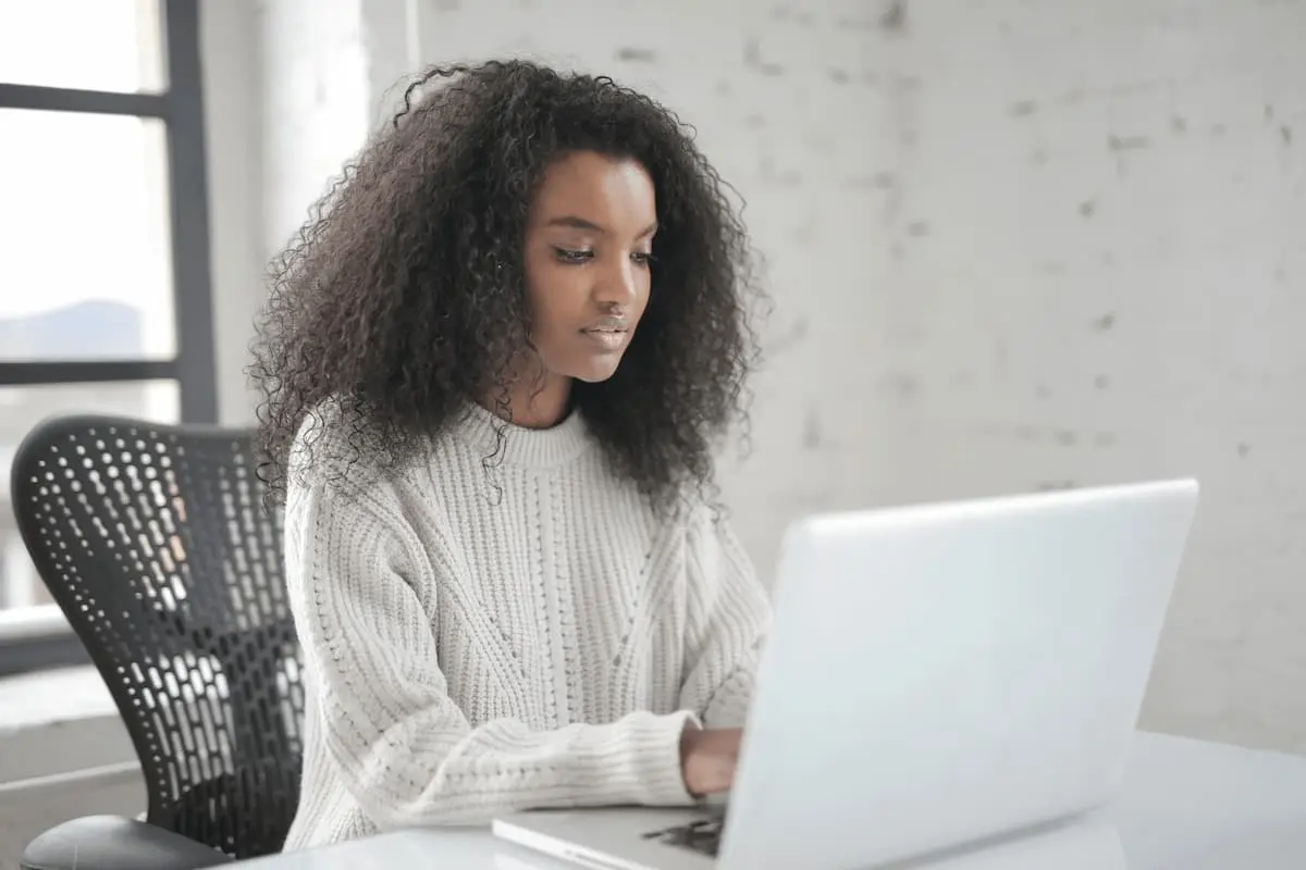Woman with curly hair wearing a gray sweater working on a laptop at a desk in a bright room.