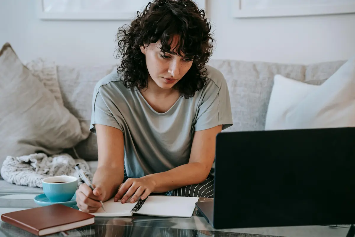 Young woman with curly hair sitting on a couch, writing in a notebook with a laptop and a cup of coffee on the table.