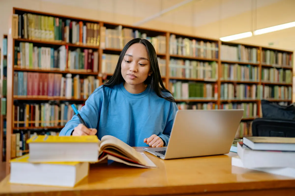 Student in blue sweater studying at a library table with open book, laptop, and stacks of books.