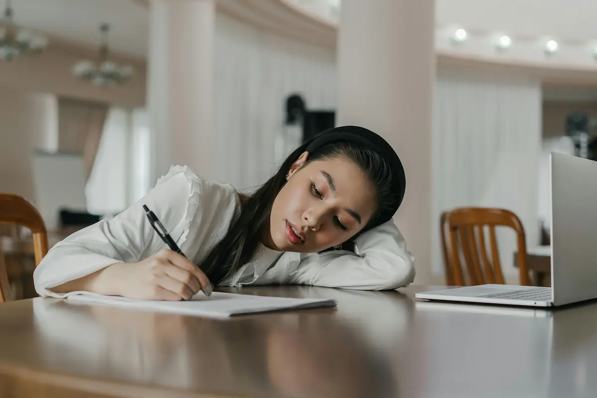 Young woman resting her head on a table while writing on paper next to an open laptop in a softly lit room.